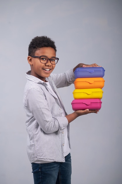 Cute smiling African American pupil in eyeglasses holding four colorful lunch boxes in his hands