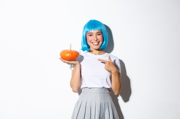 Cute smiling asian woman in blue wig pointing finger at small pumpkin, celebrating halloween, standing.