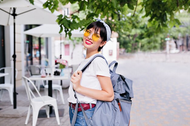 街を探索しながらバックパックを運ぶ黄色のサングラスと革ベルトを持つかわいい笑顔のブルネットの少女