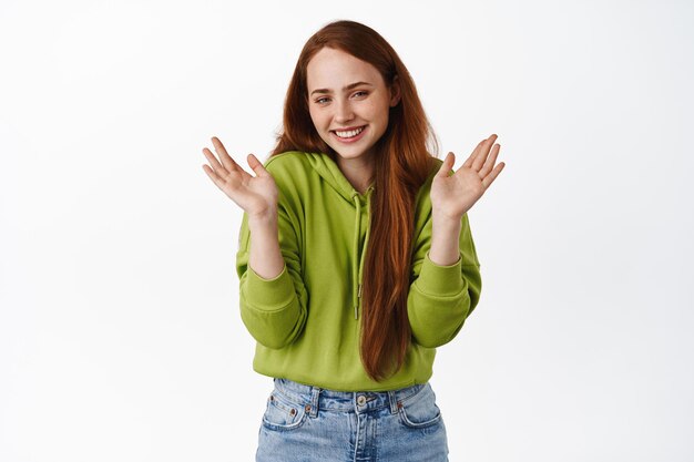 Cute smiling ginger girl, rase hands and blusing, looking happy and delighted, celebrating, standing in casual clothes against white background.