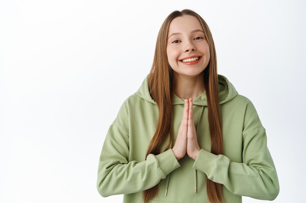 Cute smiling girl asking for favour with smile, begging pretty please, look grateful and hopeful, standing over white wall.