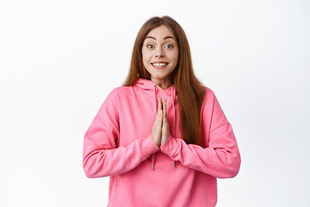 Cute smiling girl asking for help, holding hands in pray, pleading gesture and looking hopeful, standing over white wall