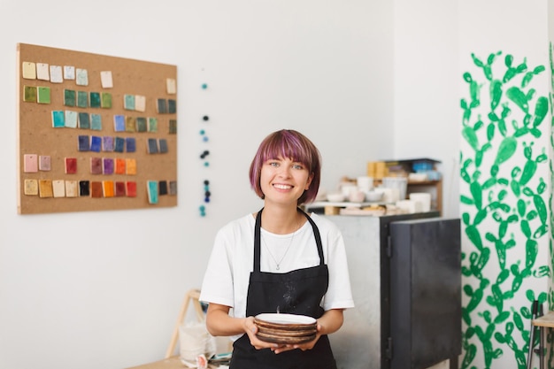 Free photo cute smiling girl in black apron and white t-shirt holding handmade plates in hands happily looking in camera at pottery studio