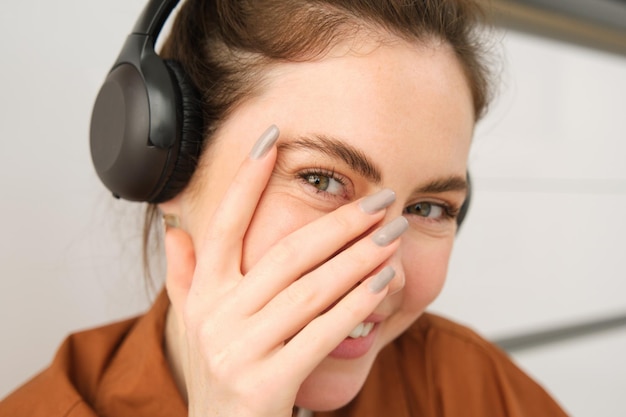 Free photo cute smiling girl blushes hides face behind hand and laughing wears wireless headphones