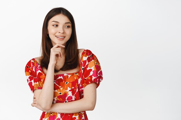 Cute smiling girl looking pleased aside gazing at logo or brand name wearing red floral dress against white background