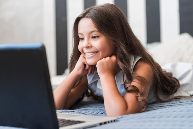 Cute smiling girl lying on bed watching video on laptop