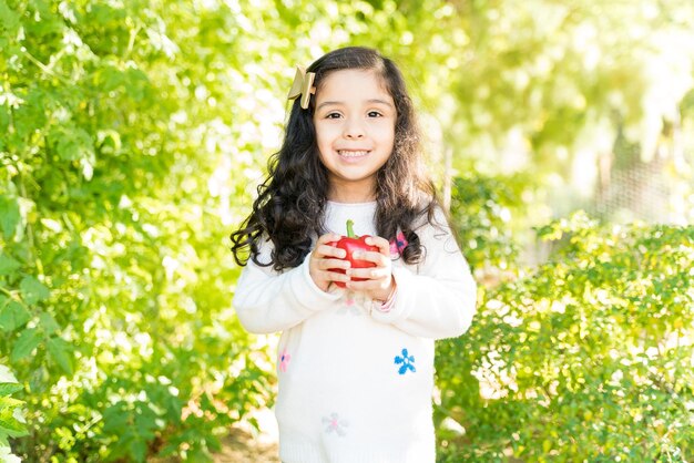 Cute smiling Hispanic girl holding fresh bell pepper while standing at farm