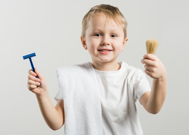 Cute smiling little boy holding razor and shaving brush standing against white background