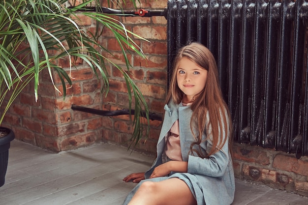 Милая улыбающаяся маленькая девочка с длинными каштановыми волосами в стильном платье сидит на деревянном полу в комнате с лофтовым интерьером.