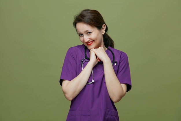 Милая улыбающаяся женщина-врач средних лет в униформе и со стетоскопом на шее держит руки вместе под подбородком и смотрит в камеру, изолированную на оливково-зеленом фоне