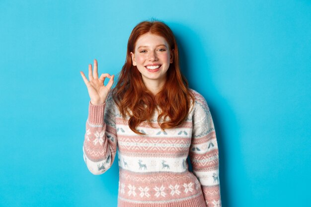 Cute smiling redhead woman showing okay sign, praising good work, well done gesture, standing over blue background.