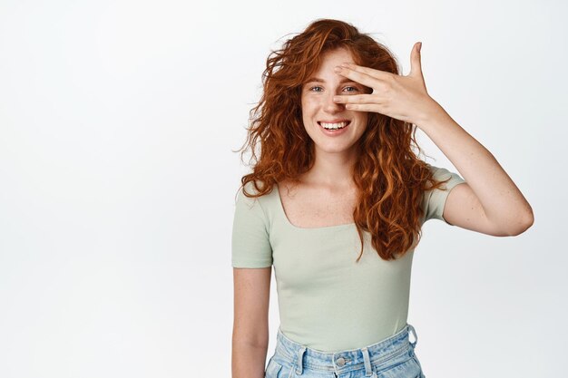 Симпатичная улыбающаяся девочка-подросток с натуральными рыжими волосами, открытыми глазами, смотрящая сквозь пальцы и выглядящая счастливой, стоя с чистой светящейся кожей в футболке на белом фоне