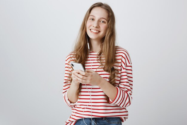 Cute smiling teenage girl using mobile phone and listening music in earphones