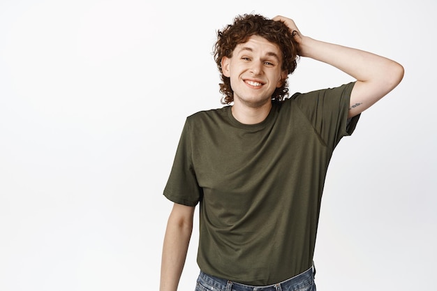 Cute smiling young man with curly hairstyle scratching head and laughing shy awkward pose standing over white background