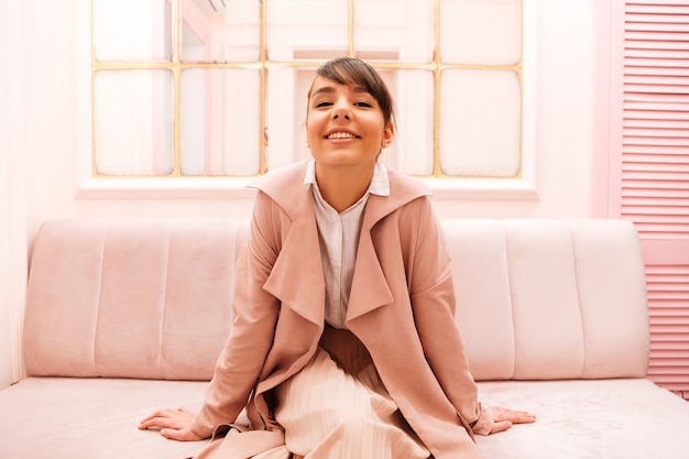 Cute smiling young woman in coat sitting on a sofa