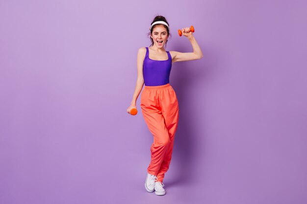 Cute sporty woman in bright fitness outfit in style of 80s with smile demonstrates exercises with dumbbells