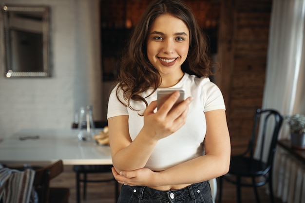 Cute student girl in white top holding cell phone, texting friends via messengaer application, making plans for weekend