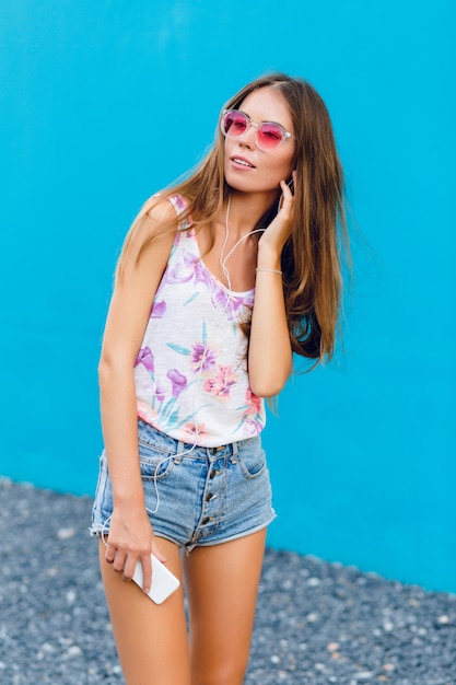 Cute stylish girl on blue background stands and listens to music on earphones on smartphone