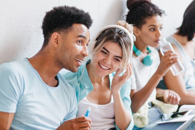 Cute stylish girl listening favorite song in headphones with friend in blue shirt while pretty young woman drinks water. Laughing students enjoying music in their free time.