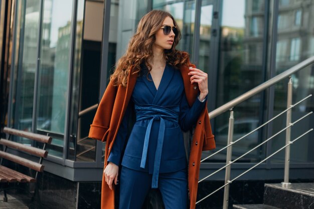 Cute stylish woman with walking in urban city business street dressed in warm brown coat and blue suit, spring autumn trendy fashion street style, wearing sunglasses