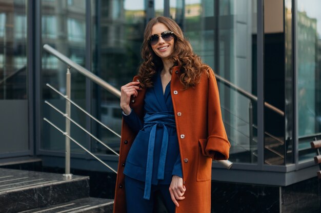 Cute stylish woman with walking in urban city business street dressed in warm brown coat and blue suit, spring autumn trendy fashion street style, wearing sunglasses