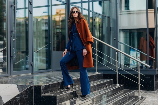 Cute stylish woman with walking in urban city business street dressed in warm brown coat and blue suit, spring autumn trendy fashion street style, wearing sunglasses