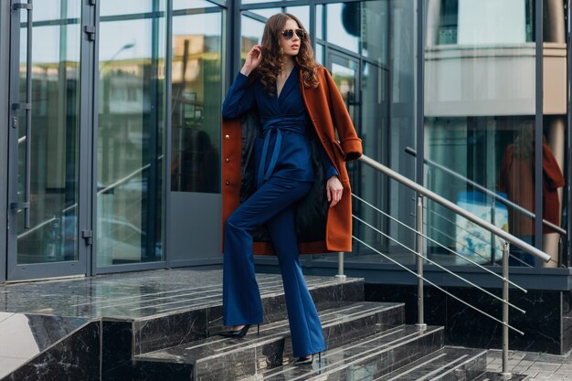 Cute stylish woman with walking in urban city business street dressed in warm brown coat and blue suit, spring autumn trendy fashion street style, wearing sunglasses