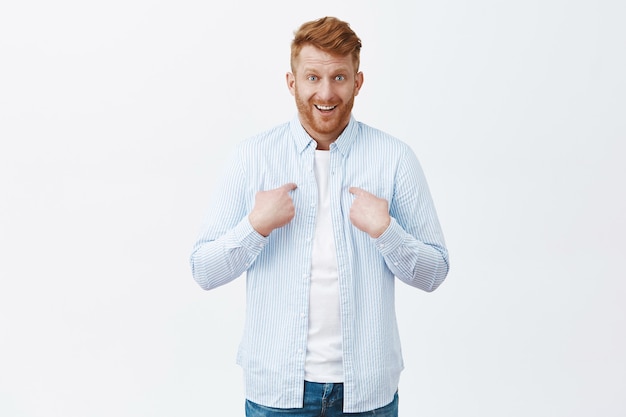 Free photo cute surprised businessman with ginger hair and bristle pointing at chest and chuckling, smirking from amazement, being picked unexpectedly, standing confused over gray wall