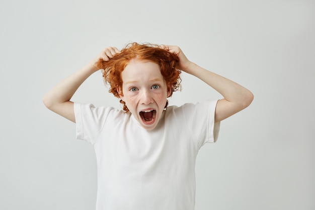 Simpatico ragazzino sorpreso con le lentiggini guardando con espressione folle e bocca aperta, tenendo i capelli con le mani.