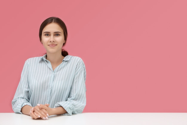 Cute talented young woman in striped shirt sitting at desk with hands clasped during job interview, her look expressing confidence and readiness.