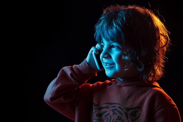 Cute talking on phone. Close up. Caucasian boy's portrait on dark studio background in neon light. Beautiful curly model. Concept of human emotions, facial expression, sales, ad, modern tech, gadgets.