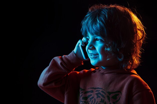 Cute talking on phone. Close up. Caucasian boy's portrait on dark studio background in neon light. Beautiful curly model. Concept of human emotions, facial expression, sales, ad, modern tech, gadgets.