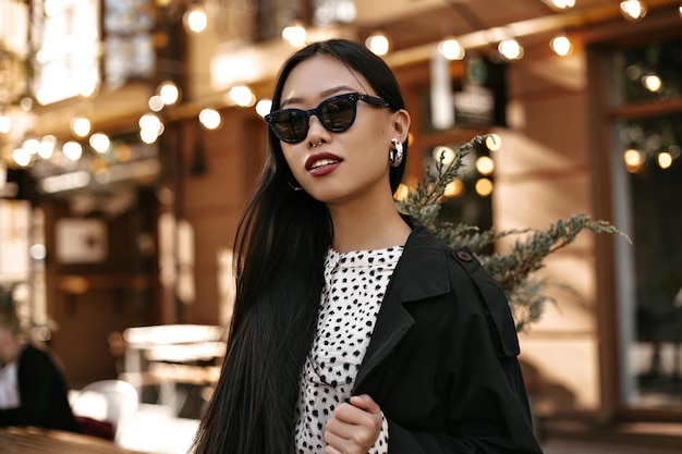 Симпатичная загорелая азиатка в черном плаще и белом платье позирует на улице Портрет брюнетки в солнцезащитных очках