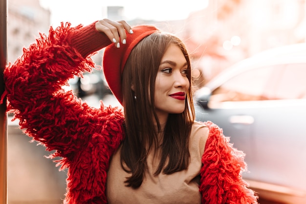 Free photo cute tanned woman in beige t-shirt, red coat and hat leaned on window and smiling against backdrop of city.