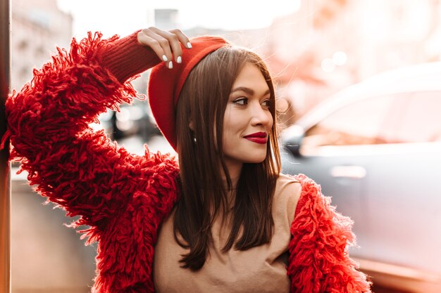 Cute tanned woman in beige T-shirt, red coat and hat leaned on window and smiling against backdrop of city.