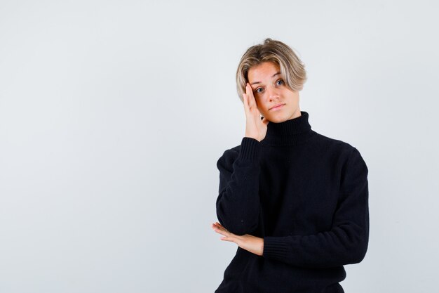 Cute teen boy in black turtleneck sweater leaning head on hand and looking pensive , front view.