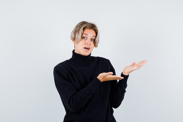 Cute teen boy doing welcome gesture in turtleneck sweater and looking surprised