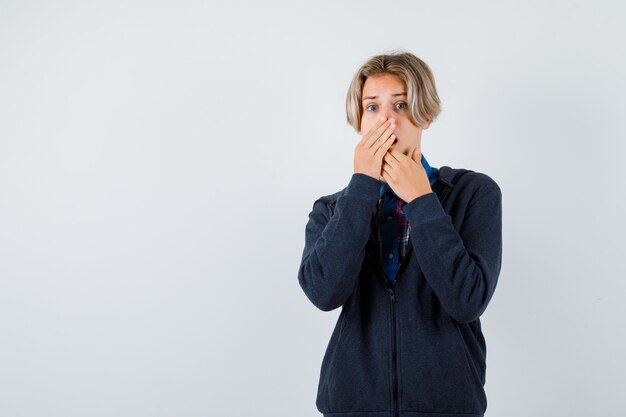 Cute teen boy in hoodie keeping hand on mouth and looking agitated , front view.