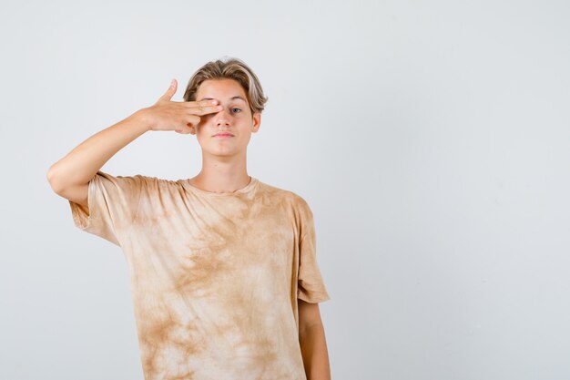 Cute teen boy keeping fingers on eye in a gun gesture in t-shirt and looking confident , front view.