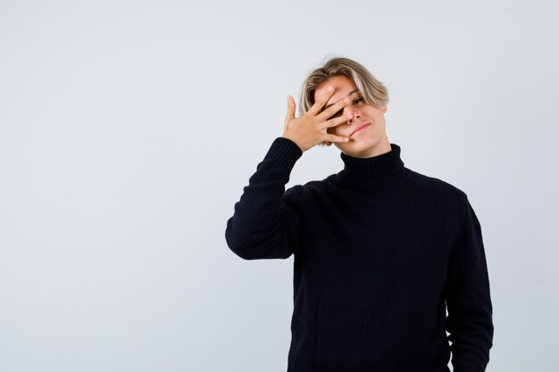 Cute teen boy keeping hand on eye in turtleneck sweater and looking dreamy