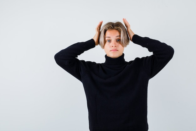 Cute teen boy keeping hands near head in turtleneck sweater and looking surprised. front view.