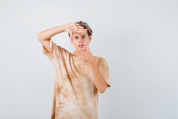 Cute teen boy looking through hands in t-shirt and looking puzzled , front view.