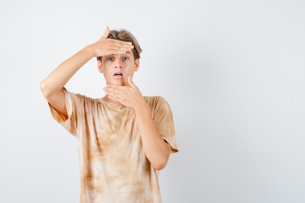 Cute teen boy looking through hands in t-shirt and looking shocked , front view.