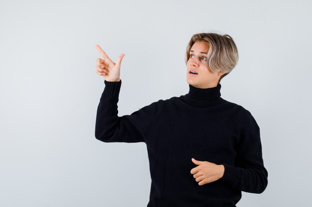 Cute teen boy pointing at upper left corner in black turtleneck sweater and looking anxious , front view.