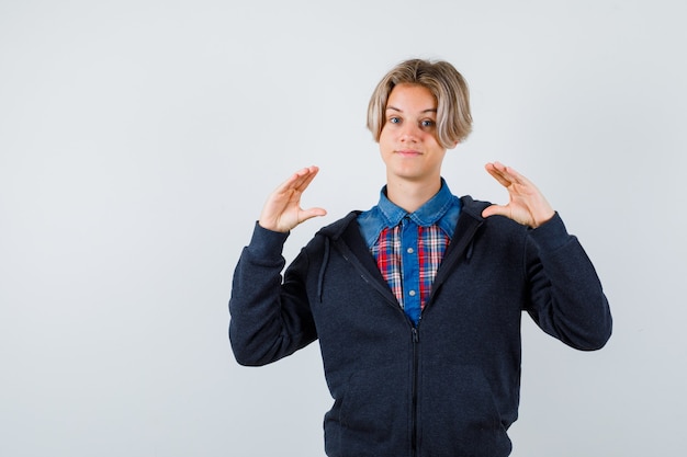 Foto gratuita ragazzo adolescente carino in camicia, felpa con cappuccio che mostra il segno della taglia e sembra allegro, vista frontale.