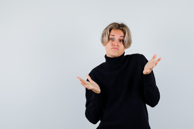 Cute teen boy showing helpless gesture in black turtleneck sweater and looking clueless , front view.