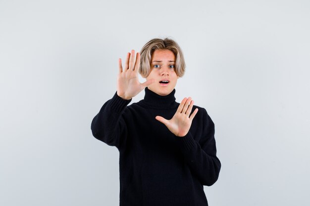 Cute teen boy showing surrender gesture in black turtleneck sweater and looking terrified. front view.
