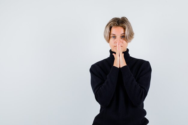 Free photo cute teen boy in turtleneck sweater showing clasped hands in pleading gesture and looking hopeful