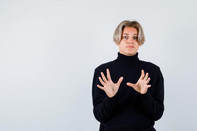 Cute teen boy in turtleneck sweater showing surrender gesture and looking frightened