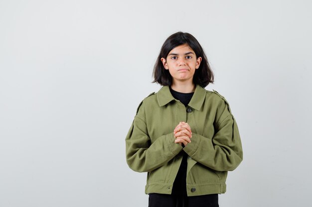 Cute teen girl in army green jacket standing with clasped hands and looking displeased , front view.